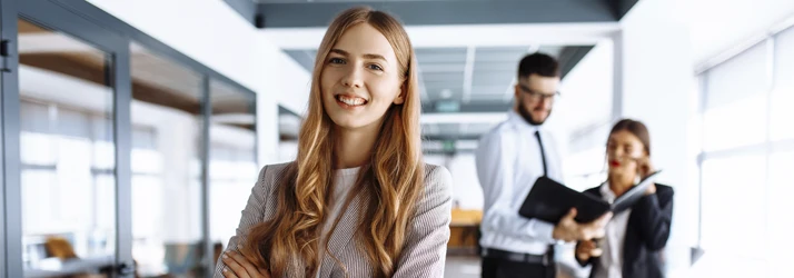 Confident professional standing in a modern office while colleagues collaborate in the background.